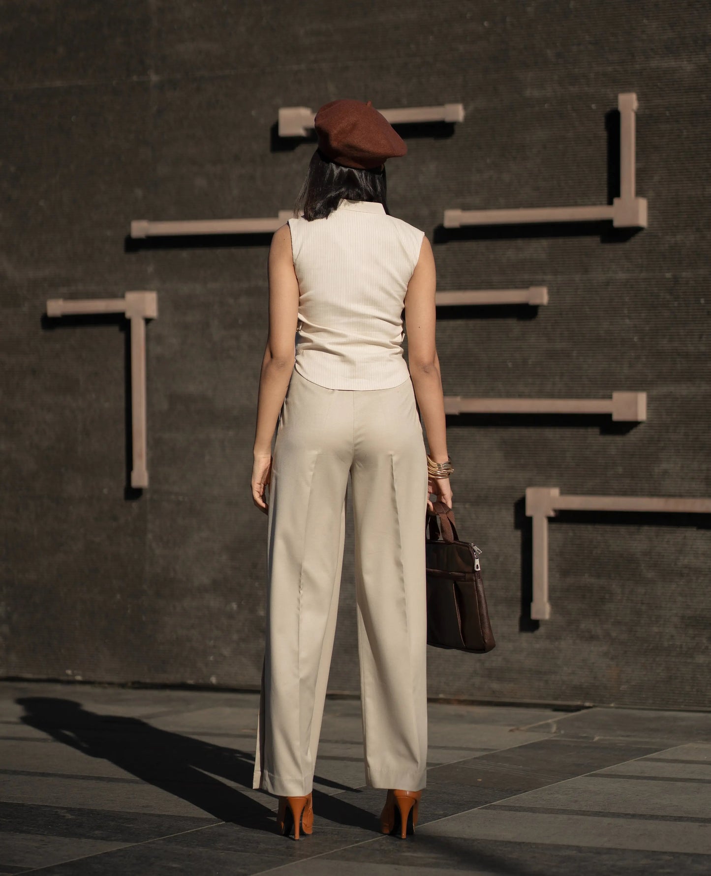 Woman in beige outfit and brown beret walking away from a dark wall with geometric shapes.