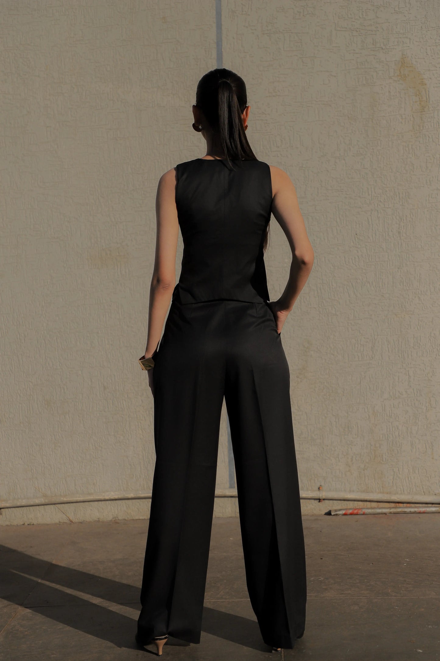 Woman wearing a black sleeveless top and wide-leg pants against a plain wall.