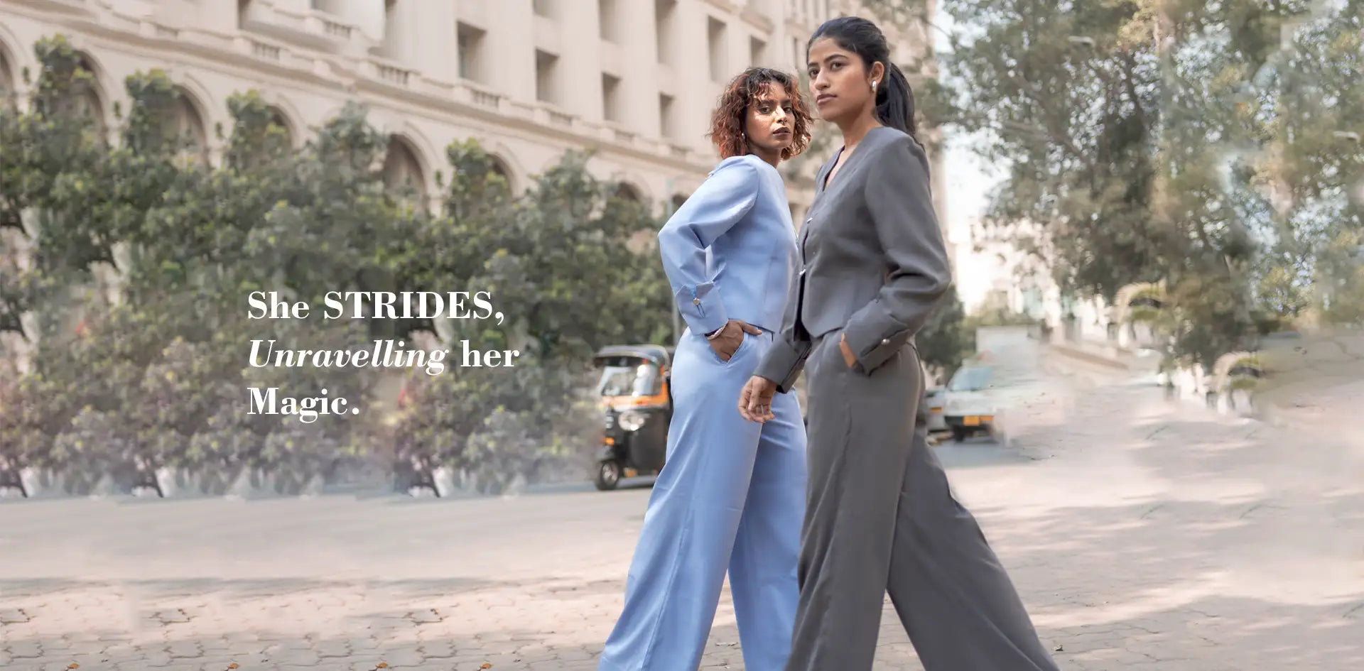 Two women walking on a street with a building in the background, featuring text 'She STRIDES, Unravelling her Magic.'