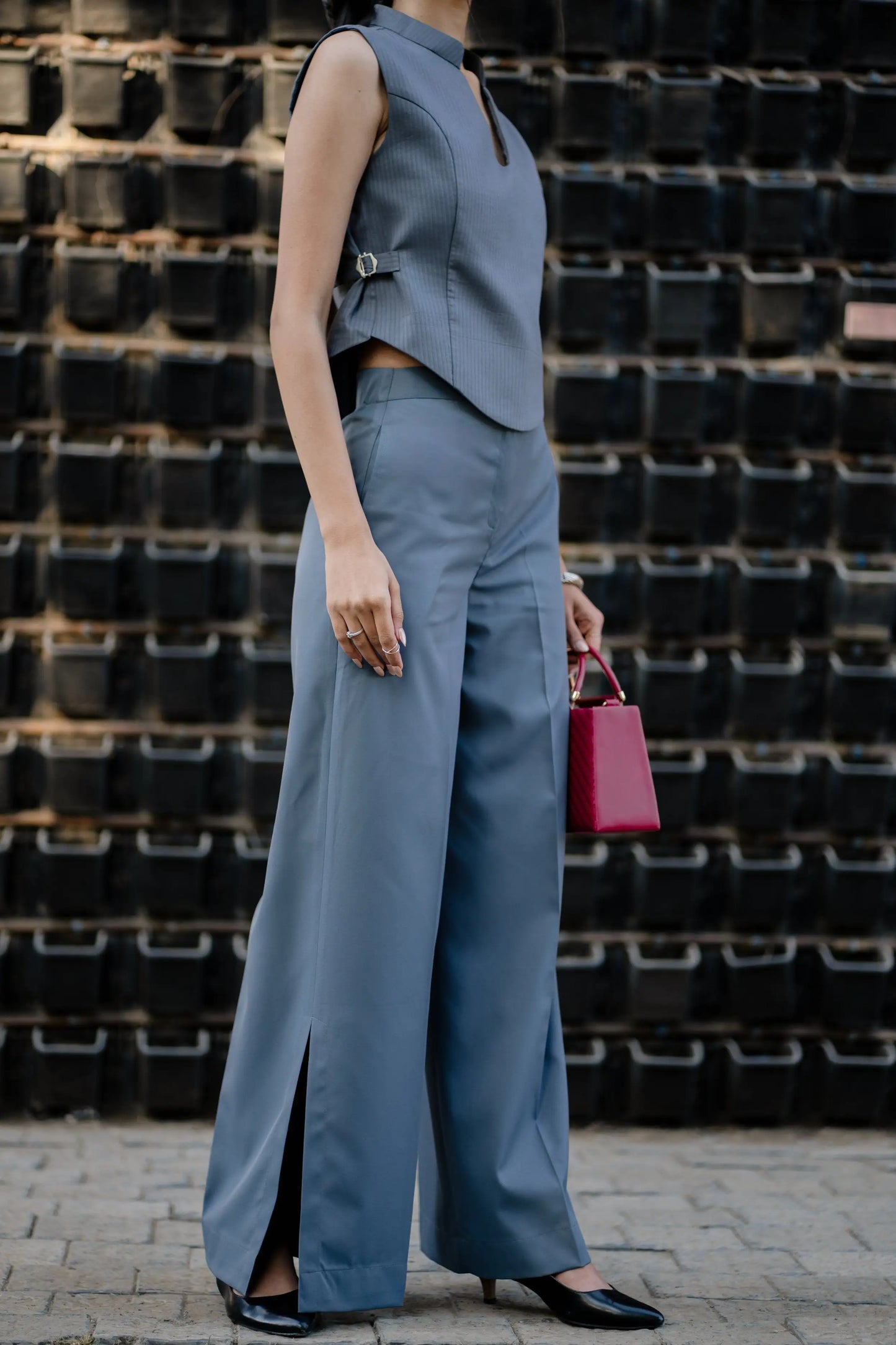 Person wearing a gray sleeveless top and wide-leg pants holding a pink handbag against a textured wall.