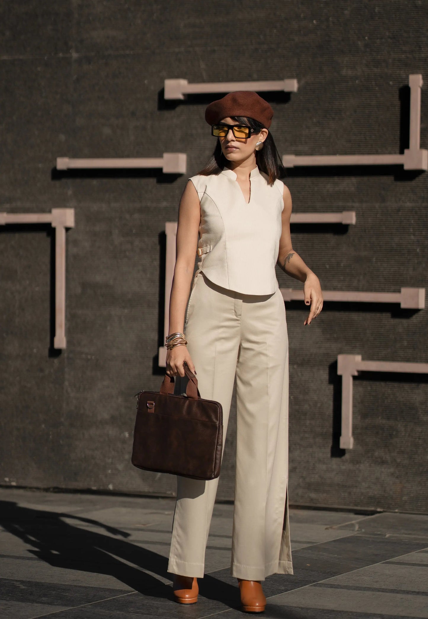 Woman in a sleeveless top and beige pants holding a brown bag against a dark background