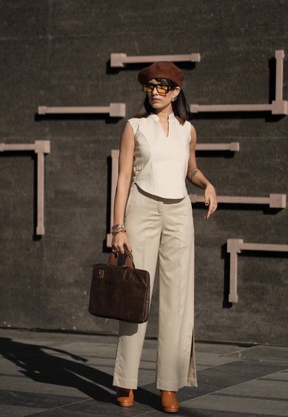 Woman in a sleeveless top and beige pants holding a brown bag against a dark background