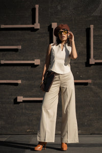 Woman in a white sleeveless top and beige pants standing against a dark wall.