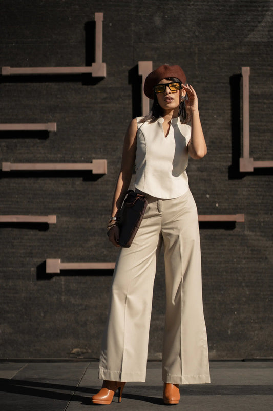 Woman in a white sleeveless top and beige pants standing against a dark wall.