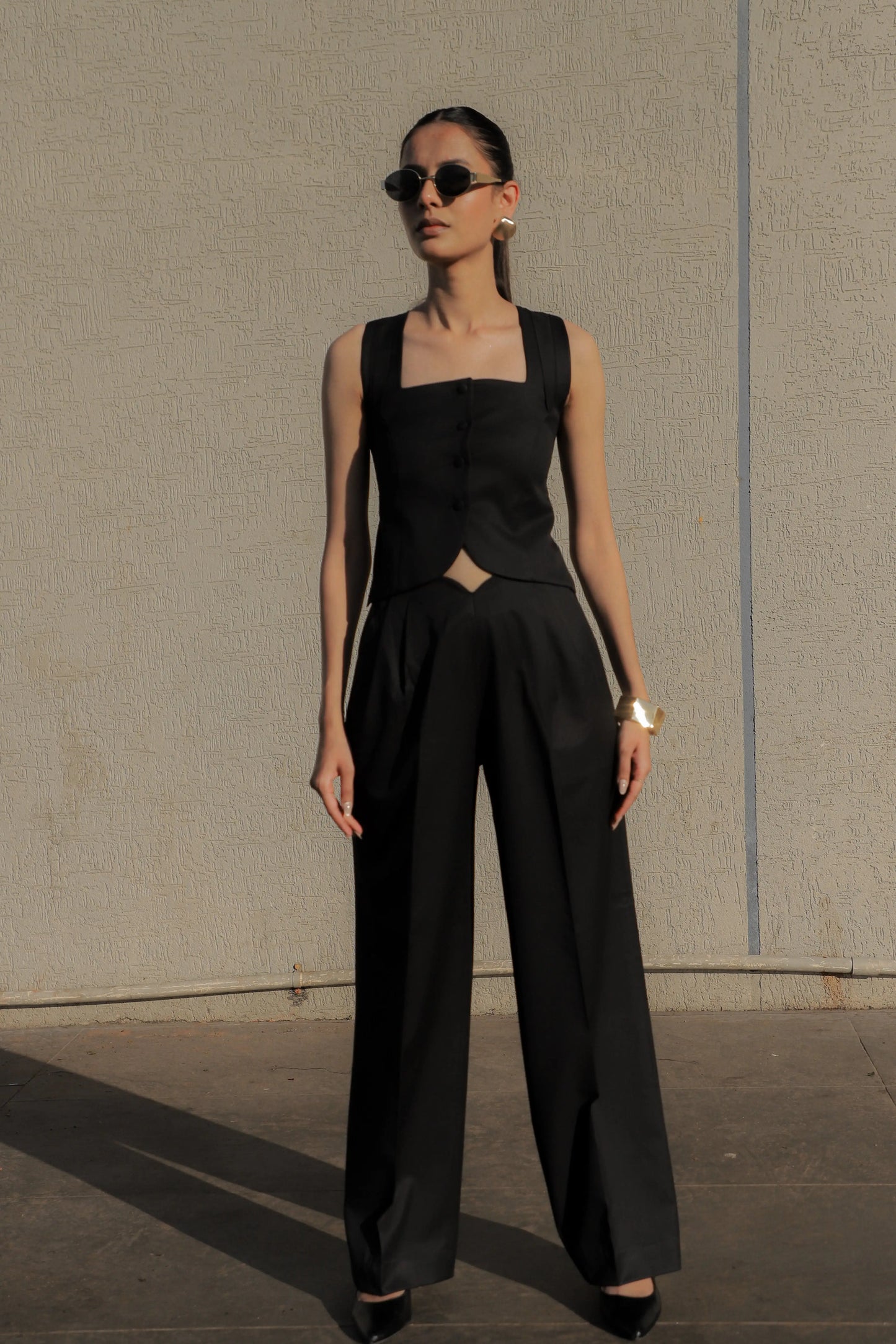 Woman wearing a black sleeveless top and high-waisted pants against a plain wall.