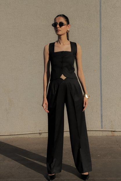 Woman wearing a black sleeveless top and high-waisted pants against a plain wall.