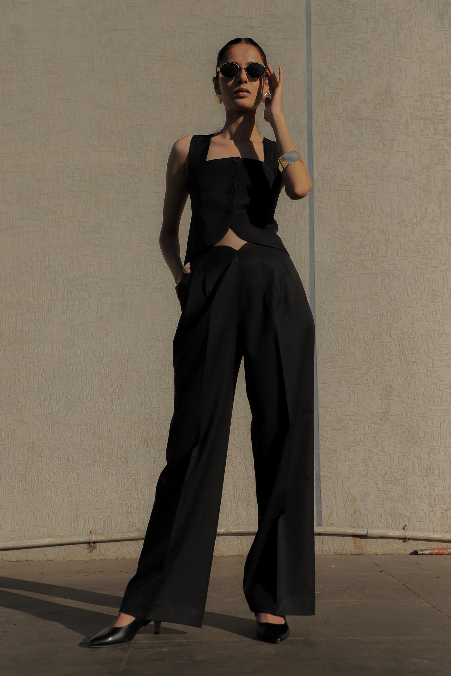 Woman wearing a black sleeveless top and wide-leg pants against a plain wall.