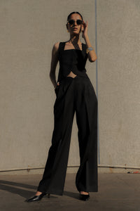 Woman wearing a black sleeveless top and wide-leg pants against a plain wall.