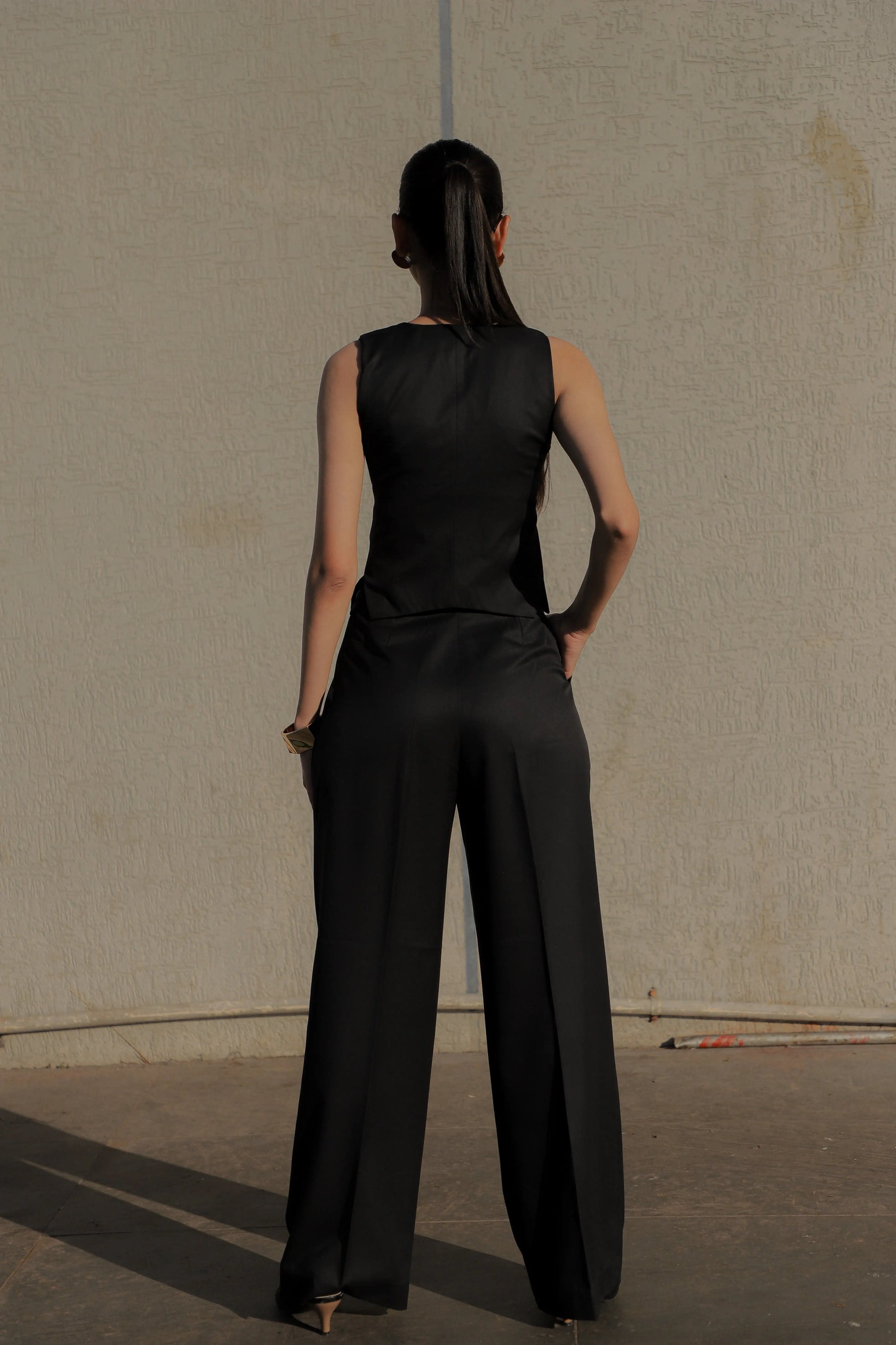 Woman wearing a black sleeveless top and wide-leg pants against a plain wall.