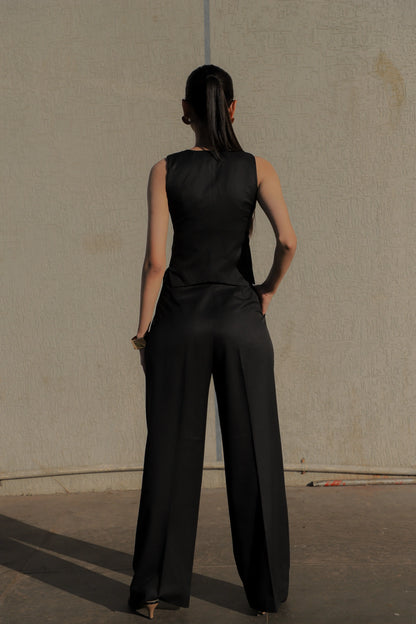 Woman wearing a black sleeveless top and wide-leg pants against a plain wall.